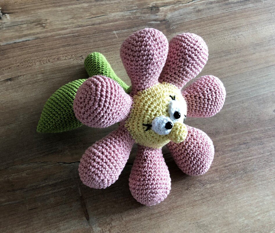 Gehäkelte Blumenrassel mit rosa Blüten, gelbem Gesicht und grünem Blatt auf Holzuntergrund