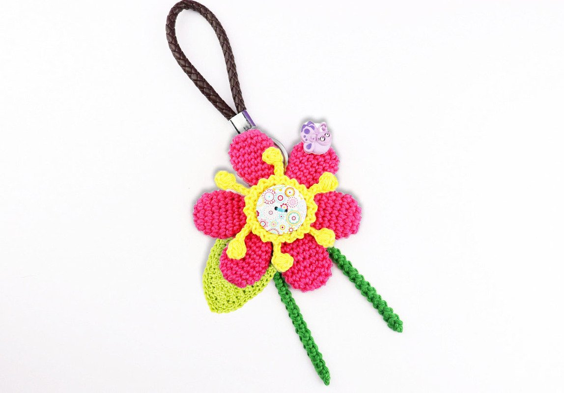 Handgehäkelter Blumen-Schlüsselanhänger in Pink und Gelb mit grünem Blatt und Schmetterlingsknopf vor weißem Hintergrund