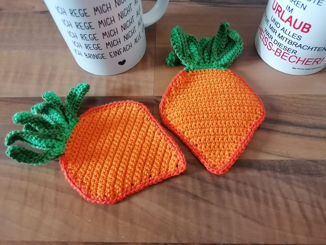 Zwei orange gehäkelte Möhren-Untersetzer mit grünen Blättern auf einem Holztisch neben Tassen