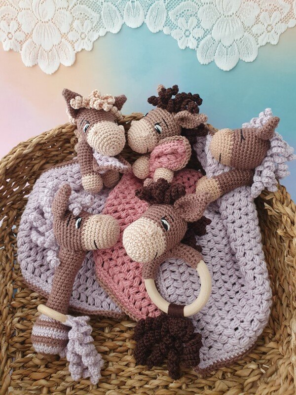 Korb mit mehreren gehäkelten Esel‑Schmusetüchern und Greiflingen, einer mit Holzring, in gedeckten Pastellfarben auf gehäkelter Decke