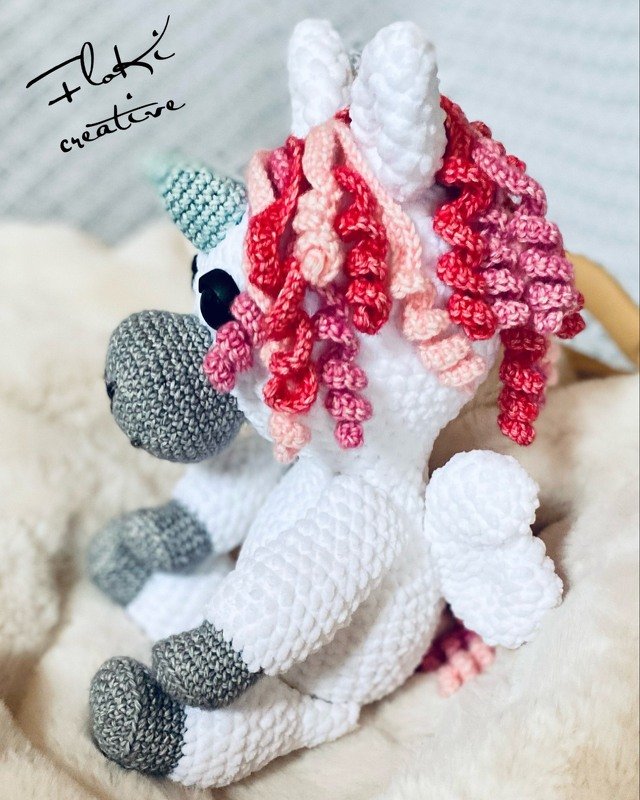 Sitzendes gehäkeltes Einhorn-Amigurumi weiß mit rosa-roter Lockenmähne, grauer Schnauze und mintfarbenem Horn