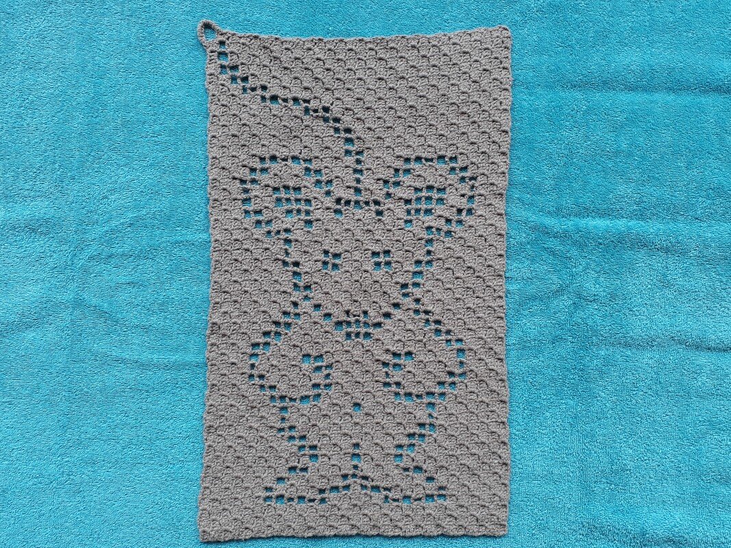 Handgehäkelter grauer Lappen mit filigranem Lochmuster und Aufhängeschlaufe auf türkisblauem Untergrund.