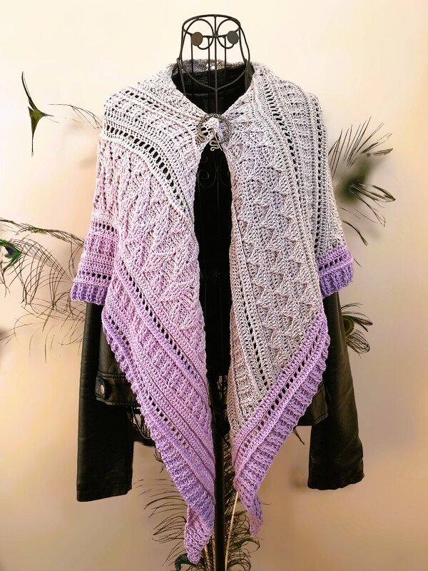 Triangular crochet shawl in a gray-to-lavender gradient draped over a black jacket on a mannequin, fastened with a shawl pin.