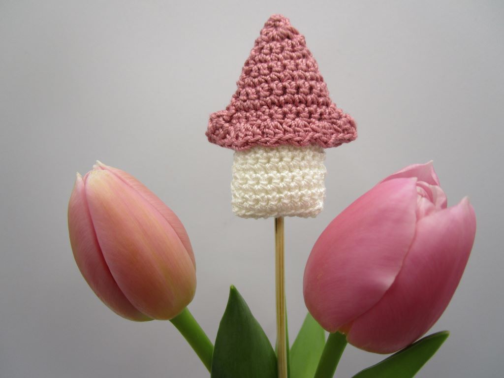 Kleiner gehäkelter Pilz mit rosa Kappe und weißem Stiel auf einem Holzstab zwischen zwei rosafarbenen Tulpen.