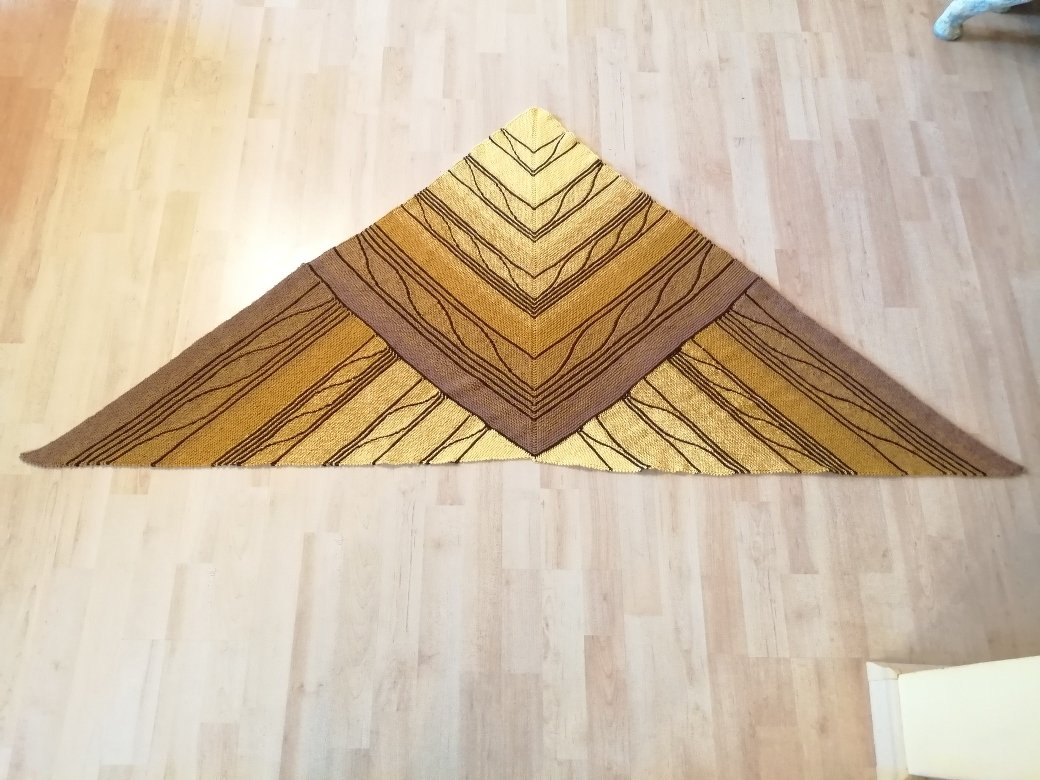 Gestricktes Dreieckstuch in Gelb- und Brauntönen mit wellenförmigen Streifen, auf hellem Holzboden ausgebreitet