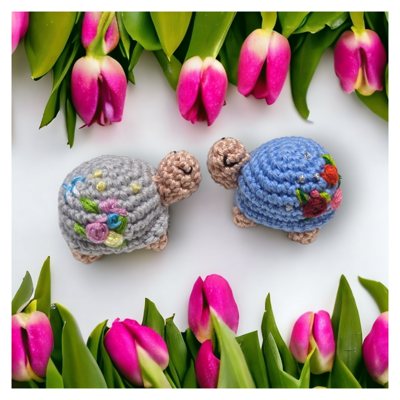 Zwei Mini‑Amigurumi‑Schildkröten in Grau und Blau mit gestickten Blumen auf den Panzern, liegend auf weißem Hintergrund zwischen Tulpen