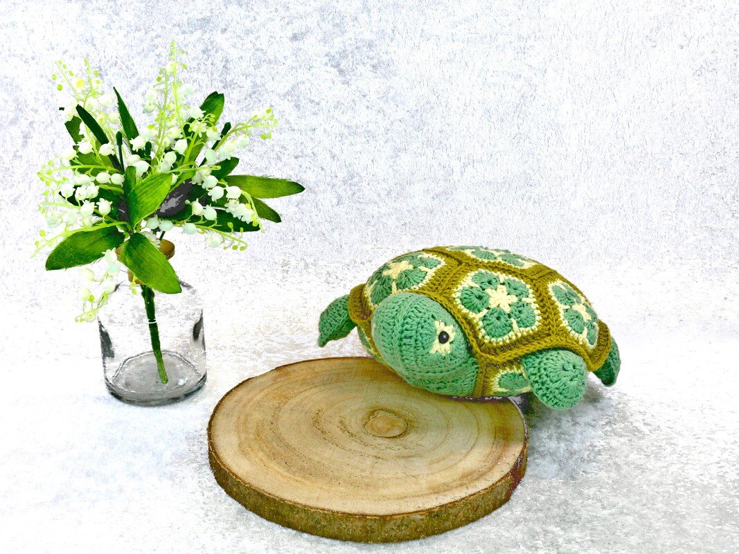 Crocheted green amigurumi turtle with African flower granny motif shell on a wood slice next to a glass vase of small white flowers