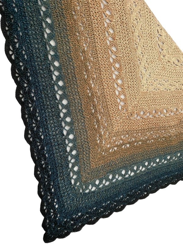 Close-up of an ombre crochet triangle shawl with a gradient from light brown to dark teal, showing a lacy openwork stitch pattern and scalloped edge.