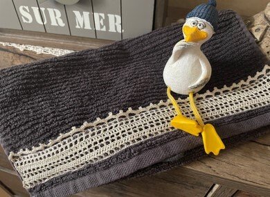 Dunkelgraues Frotteehandtuch mit cremefarbener gehäkelter Spitzenbordüre und dekorativer Möwenfigur mit blauer Mütze.