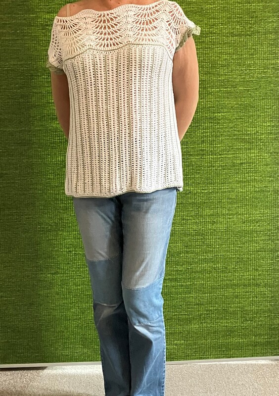 Weißes, schulterfreies Häkelshirt mit Fächermuster am Oberteil und grünen Kanten, getragen zu Jeans vor grünem Hintergrund.