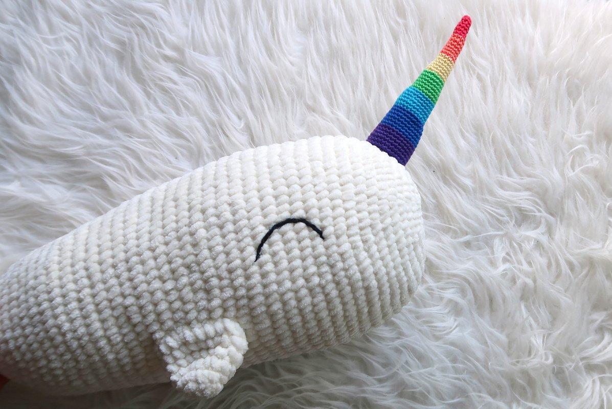 Weißes, gehäkeltes Narwal-Amigurumi mit gesticktem Gesicht und Regenbogenhorn auf flauschigem Untergrund