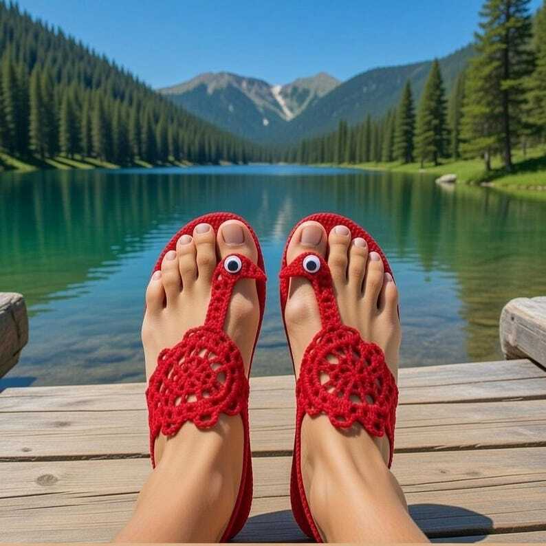 Füße in roten gehäkelten Sandalen mit Vogelapplikationen und Wackelaugen auf einem Holzsteg vor einem Bergsee.