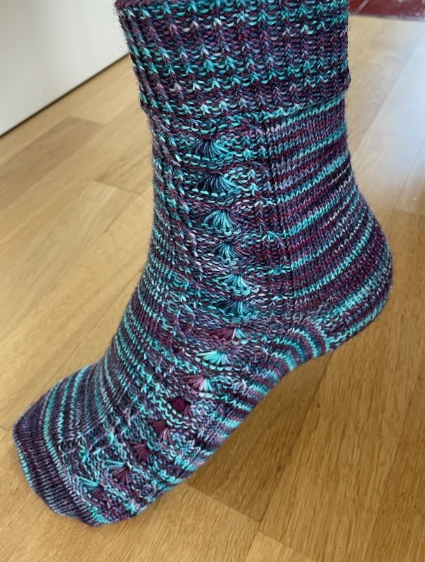 Handgestrickte Socke in Türkis, Lila und Weiß mit Noppen- und Zopfmuster.