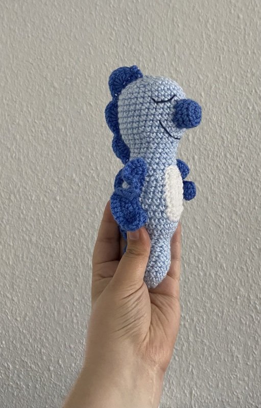 Hand hält hellblaues gehäkeltes Seepferdchen-Amigurumi mit dunkelblauen Flossen und weißem Bauch