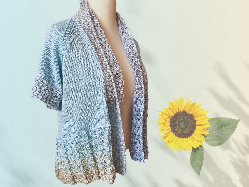 Kurzärmelige, offene Damen-Strickjacke mit Ombre-Farbverlauf und Ajourmuster an Saum und Kragen, präsentiert auf einer Büste neben einer Sonnenblume