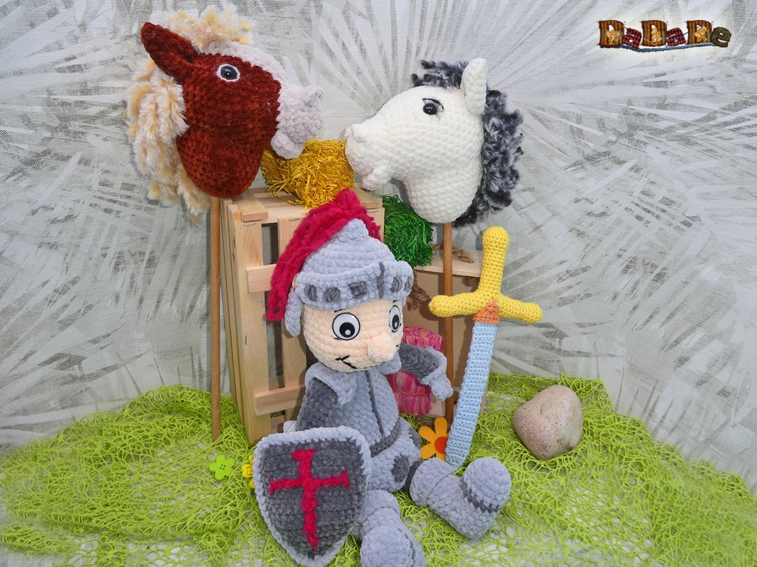 Gehäkelter Amigurumi-Ritter mit rotem Helmputz, Schild mit rotem Kreuz, hellblauem Schwert und zwei Steckenpferden