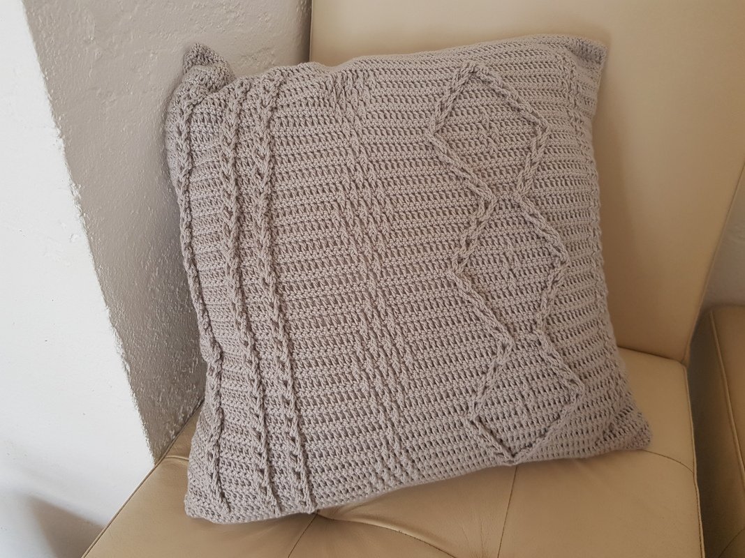 Beiges gehäkeltes Kissen mit Zopf- und Rautenmuster auf hellem Sofa