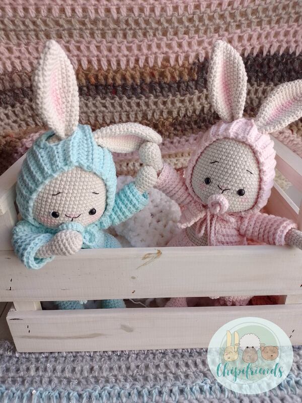 Zwei gehäkelte Amigurumi-Hasen in blau und rosa mit Kapuzenohren, sitzend in einer Holzkiste