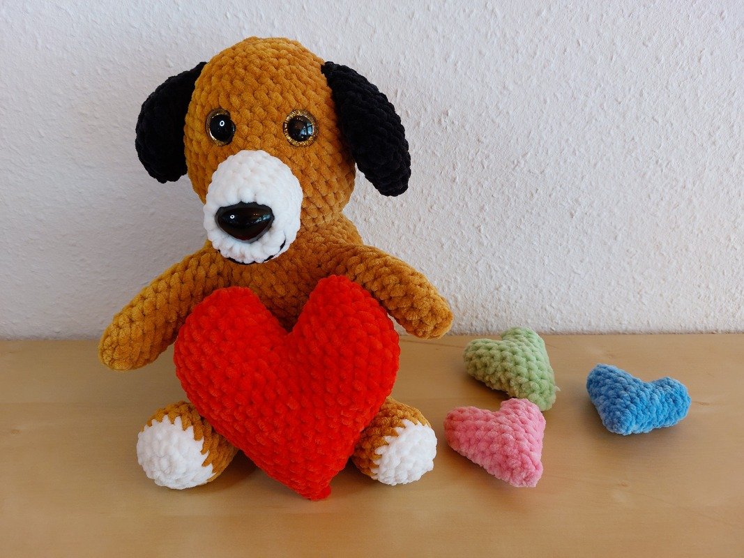Gehäkelter Amigurumi-Hund in Braun mit schwarzen Ohren, der ein großes rotes Herz hält; daneben kleine Häkelherzen