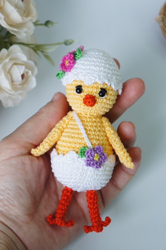 Hand holding small yellow crochet chick in a white eggshell hat and skirt, with orange legs and a tiny shoulder bag with a purple flower