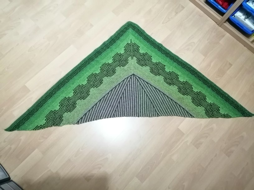 Gestricktes grünes Dreieckstuch mit keltischem Knotenmuster und Streifen, flach auf hellem Holzboden