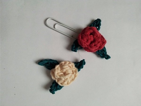Zwei gehäkelte Blumen-Lesezeichen, eine rote und eine beige, jeweils mit grünen Blättern an Büroklammern auf hellem Hintergrund.