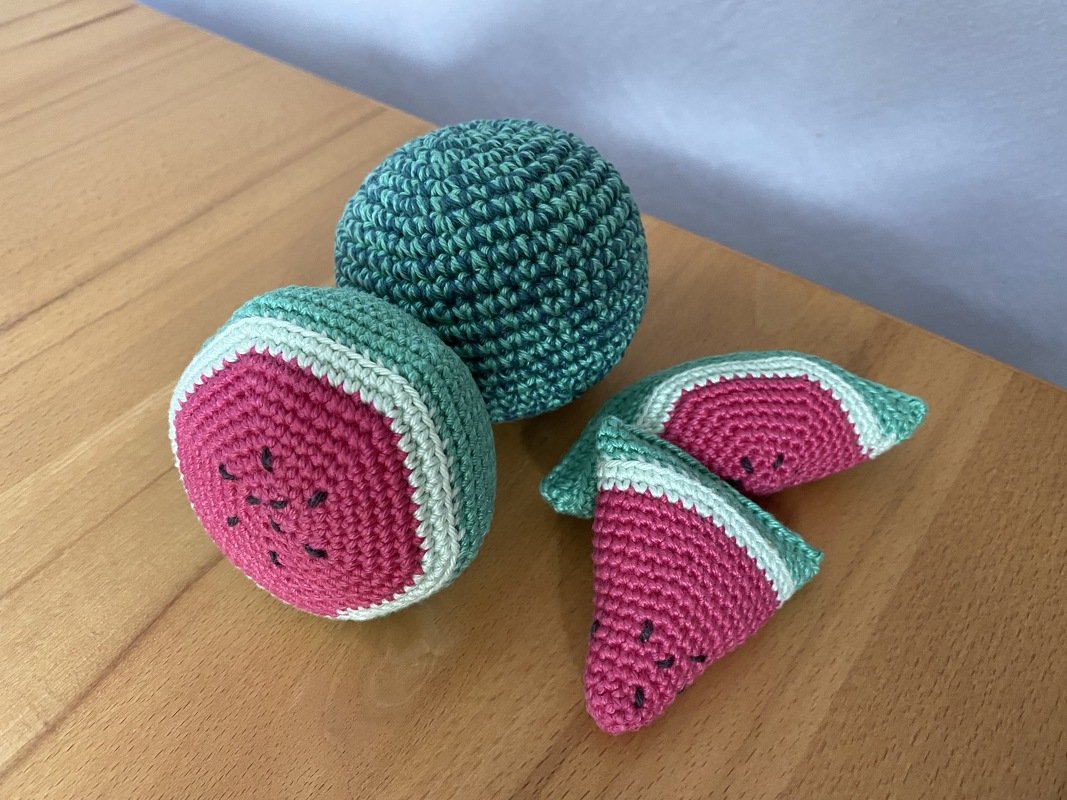 Nahaufnahme gehäkelter Wassermelonen: eine ganze Kugel, eine halbe Melone und zwei Melonenstücke in Grün, Weiß und Pink mit schwarzen Kernen auf Holz