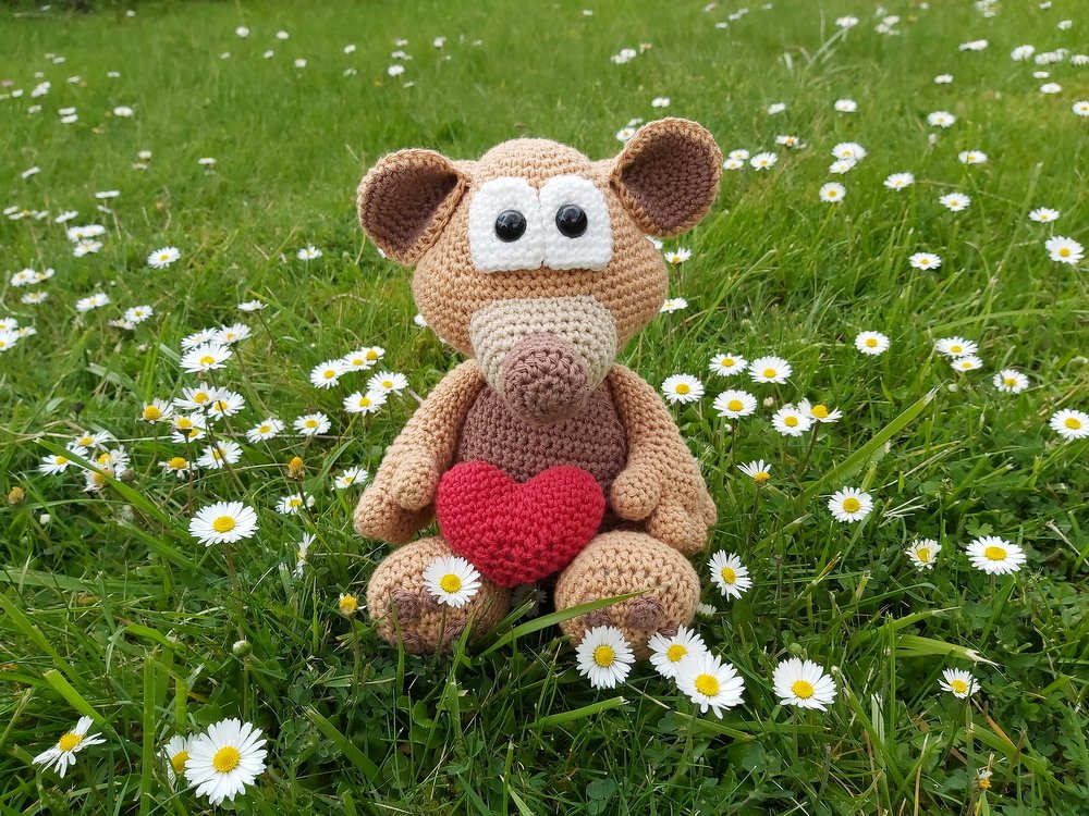 Gehäkelte Amigurumi-Maus in Beige und Braun mit rotem Herz, sitzend im Gras zwischen Gänseblümchen