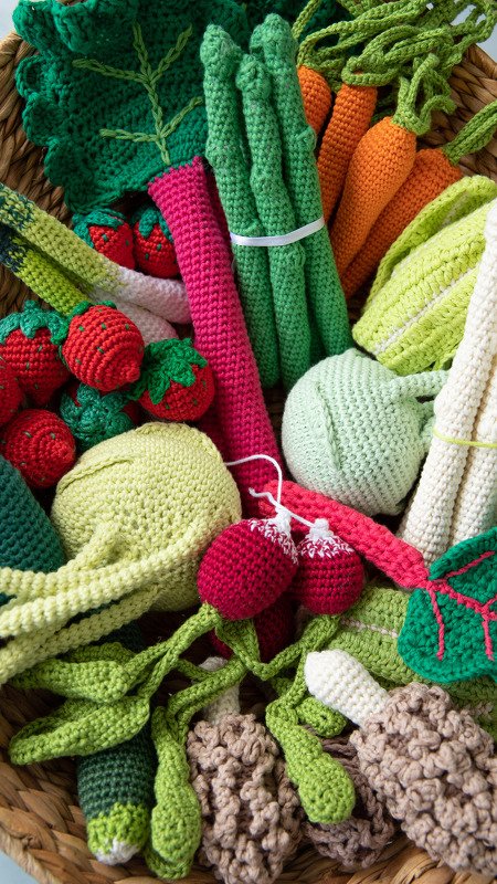 Blick von oben in einen Weidenkorb, gefüllt mit farbig gehäkeltem Spielzeug-Obst und -Gemüse wie Spargel, Karotten, Erdbeeren, Radieschen und Pilzen.