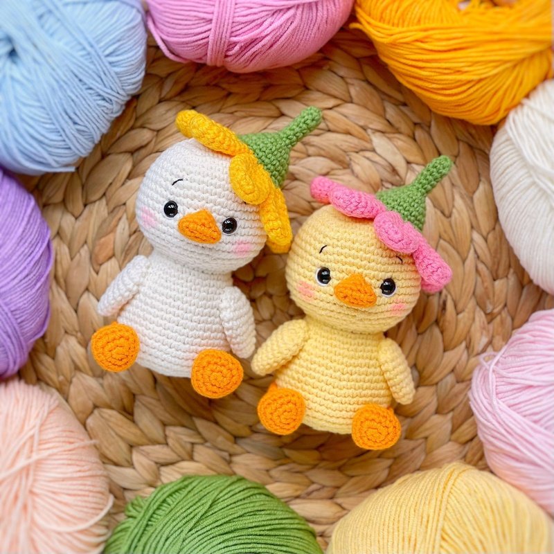Two crocheted amigurumi chicks with orange beaks and feet, wearing small flower-style hats, on a woven mat surrounded by colorful yarn balls.