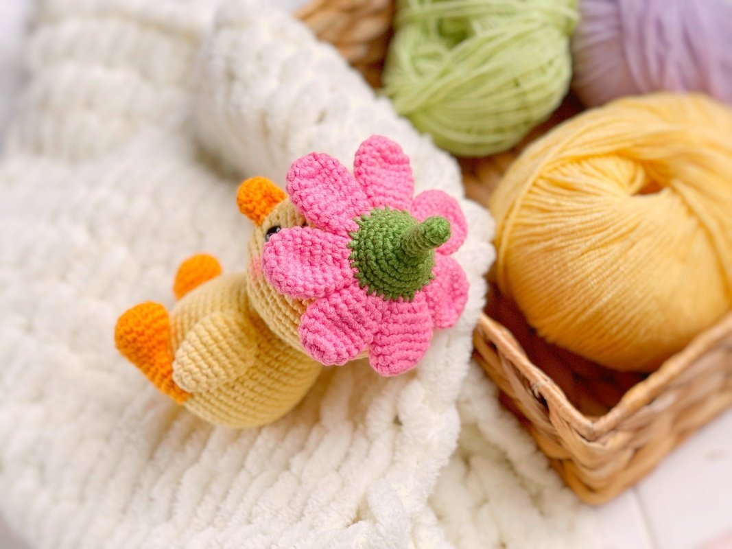 Yellow crocheted amigurumi chick with a pink flower on a white blanket beside a basket of yarn.