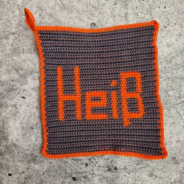 Gehäkelter grauer Topflappen mit orangefarbener Umrandung und dem Schriftzug „Heiß“, mit Aufhängeschlaufe