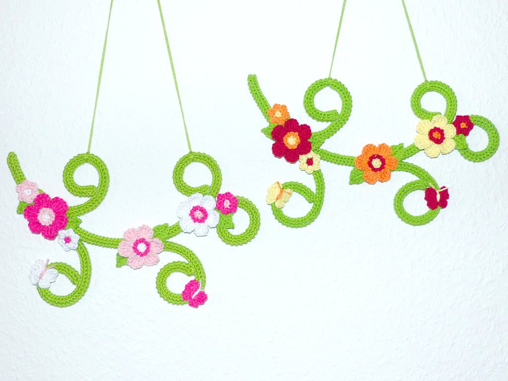 Zwei grüne gehäkelte Ranken mit bunten Blumen und kleinen Schmetterlingen vor weißer Wand