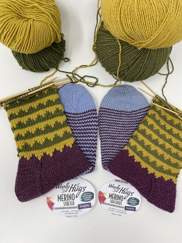 Zwei Paar farbige handgestrickte Socken mit Streifen- und Zackenmuster, Wollknäuel und Stricknadeln auf hellem Hintergrund