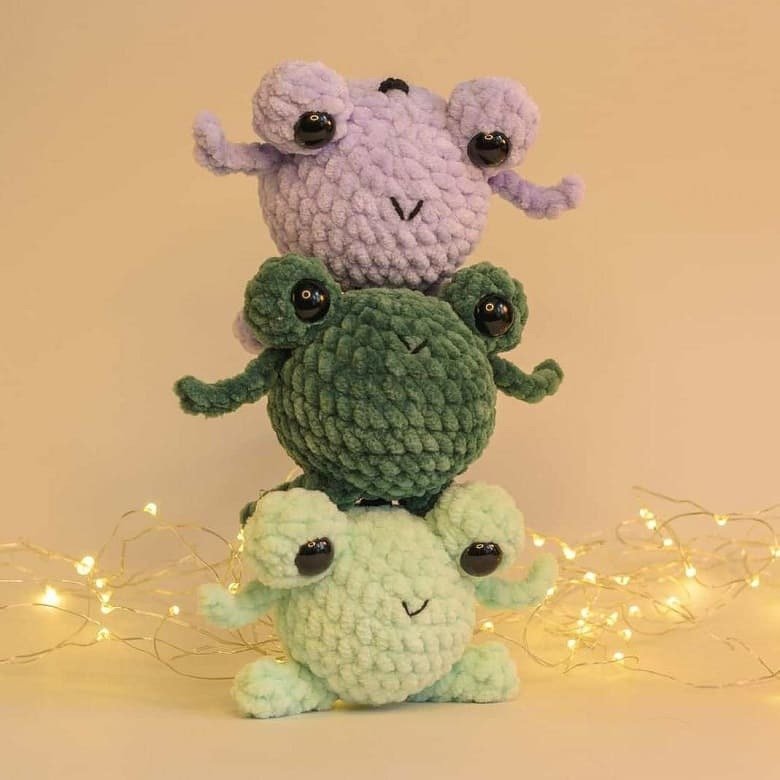 Drei gestapelte gehäkelte Frosch-Amigurumi in Pastell- und Grüntönen vor neutralem Hintergrund mit Lichterkette