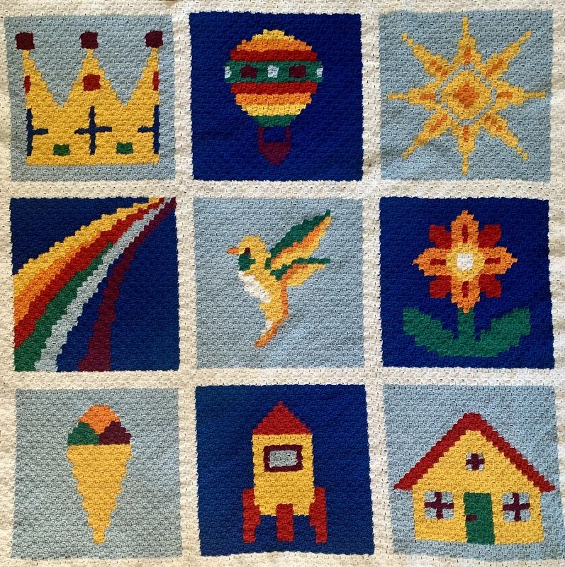 Gehäkelte Babydecke mit neun Quadraten und bunten Pixelmotiven wie Krone, Heißluftballon und Rakete.