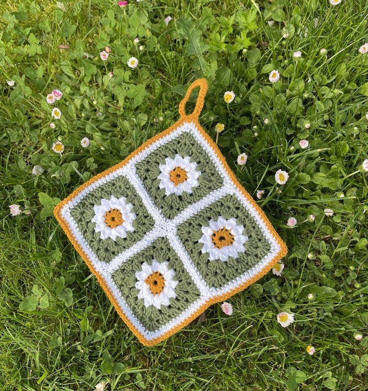 Quadratischer gehäkelter Topflappen aus vier grünen Granny‑Squares mit weißen Gänseblümchen, gelbem Zentrum und orangefarbener Umrandung, auf Gras liegend