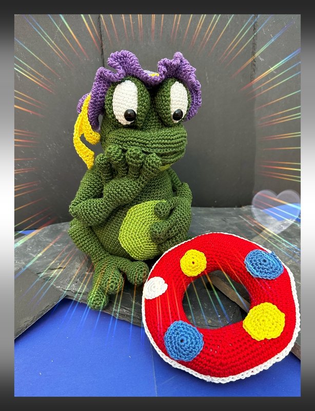 Gehäkelter grüner Frosch-Amigurumi mit lila Kopfschmuck und rotem Rettungsring auf dunklem Hintergrund.
