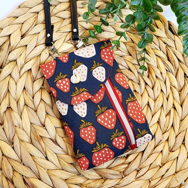 Dunkelblaue Stofftasche mit rotem und weißem Erdbeermuster, Umschlagklappe mit Druckknopf und Reißverschlussfach auf geflochtener Unterlage mit grünem Blattwerk.