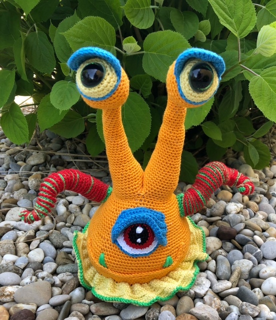 Orangefarbenes Häkelmonster (Amigurumi) mit zwei Stielaugen, Bauchauge, rot-grün gestreiften Armen und gelb gewelltem Standfuß auf Kieselsteinen.
