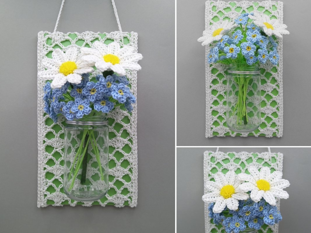 White crocheted daisies and small blue crocheted flowers in a glass jar attached to a white lacy crochet panel with green backing, hanging on a wall.