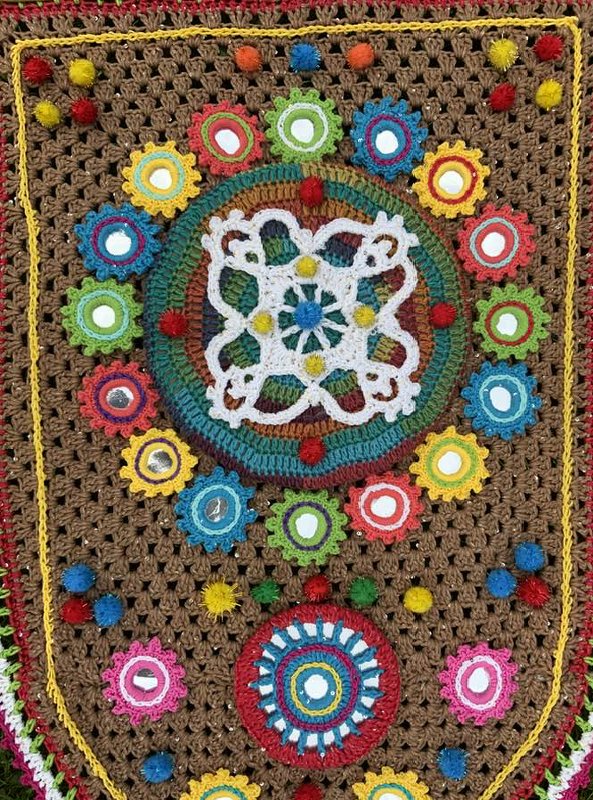 Nahaufnahme eines bunten gehäkelten Wandbehangs auf braunem Grund mit Mandalas, kleinen Spiegeln und Pompons.