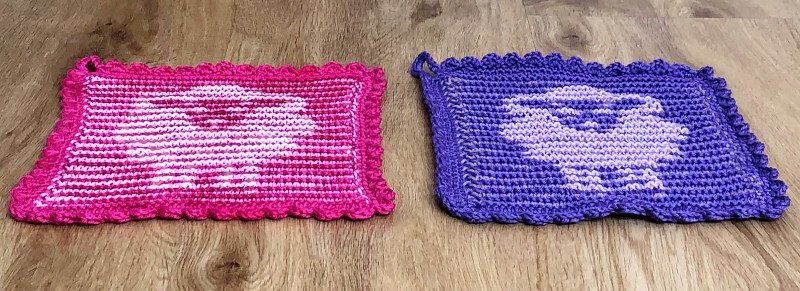 Zwei gestrickte Topflappen in Pink und Violett mit hellem Herzmuster und gewelltem Rand auf Holzoberfläche