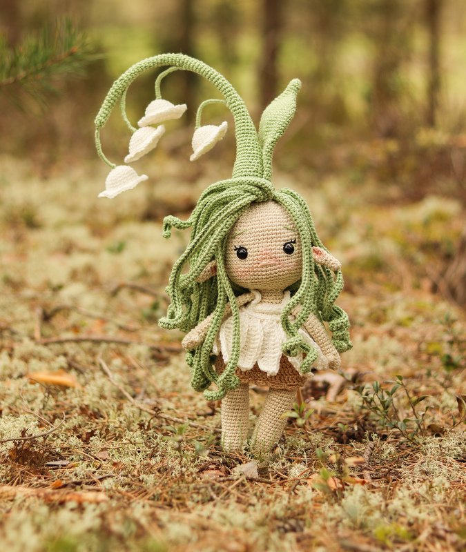 Gehäkelte Amigurumi-Puppe als Maiglöckchen-Fee mit grünen Haaren und Blütenkopfschmuck auf moosigem Waldboden