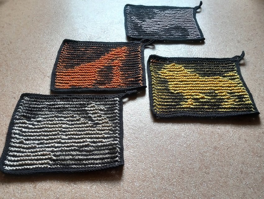 Vier gestrickte Topflappen mit kontrastreichen Tierumrissen in Orange, Gelb, Hellgrau und Beige auf dunklem Grund, alle mit dunklem Rand und Aufhängeschlaufen.