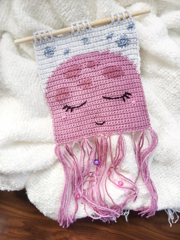 Gehäkeltes Wandbild in Form einer rosa Qualle mit geschlossenen Augen und lila Fransen, aufgehängt an einem Holzstab.