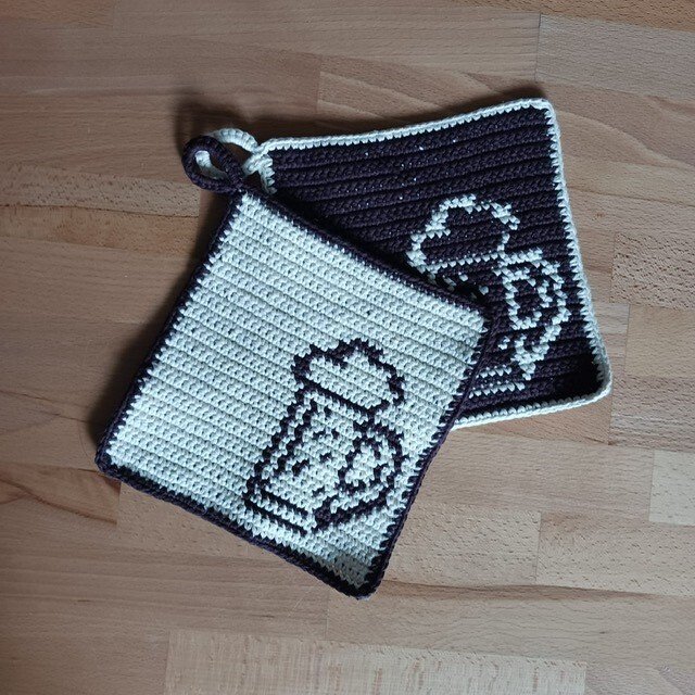Zwei quadratische gehäkelte Topflappen in Creme und Dunkelbraun mit Bierkrug-Motiv und Aufhängeschlaufe auf Holzuntergrund