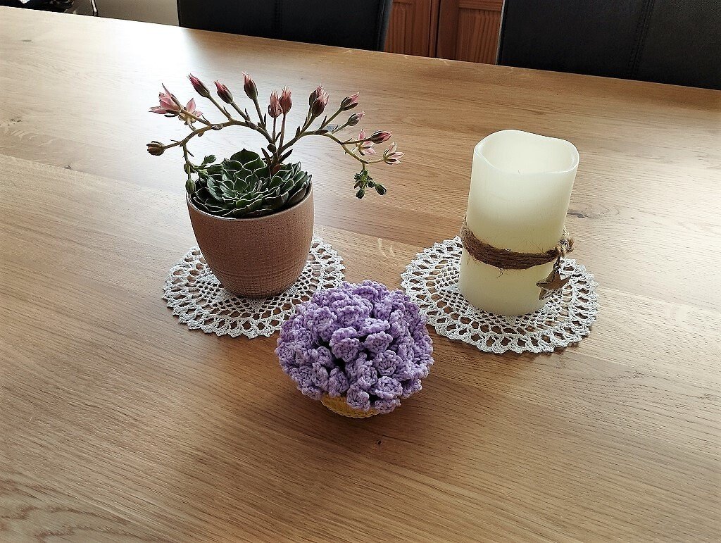 Sukkulente, Kerze und lila Häkelblüte auf weißen Spitzenuntersetzern auf Holztisch.