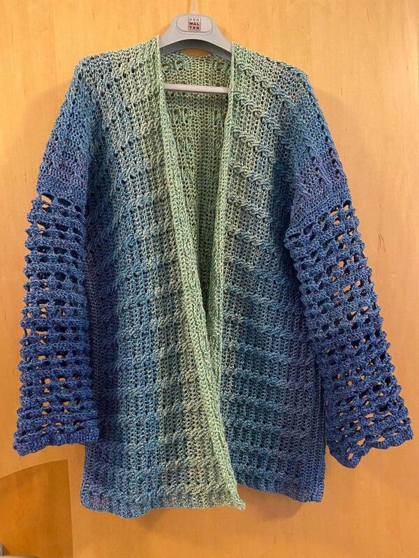 Gehäkelte Damenjacke in Blau und Grün mit Lochmuster-Ärmeln an einem Kleiderbügel vor hellem Holz