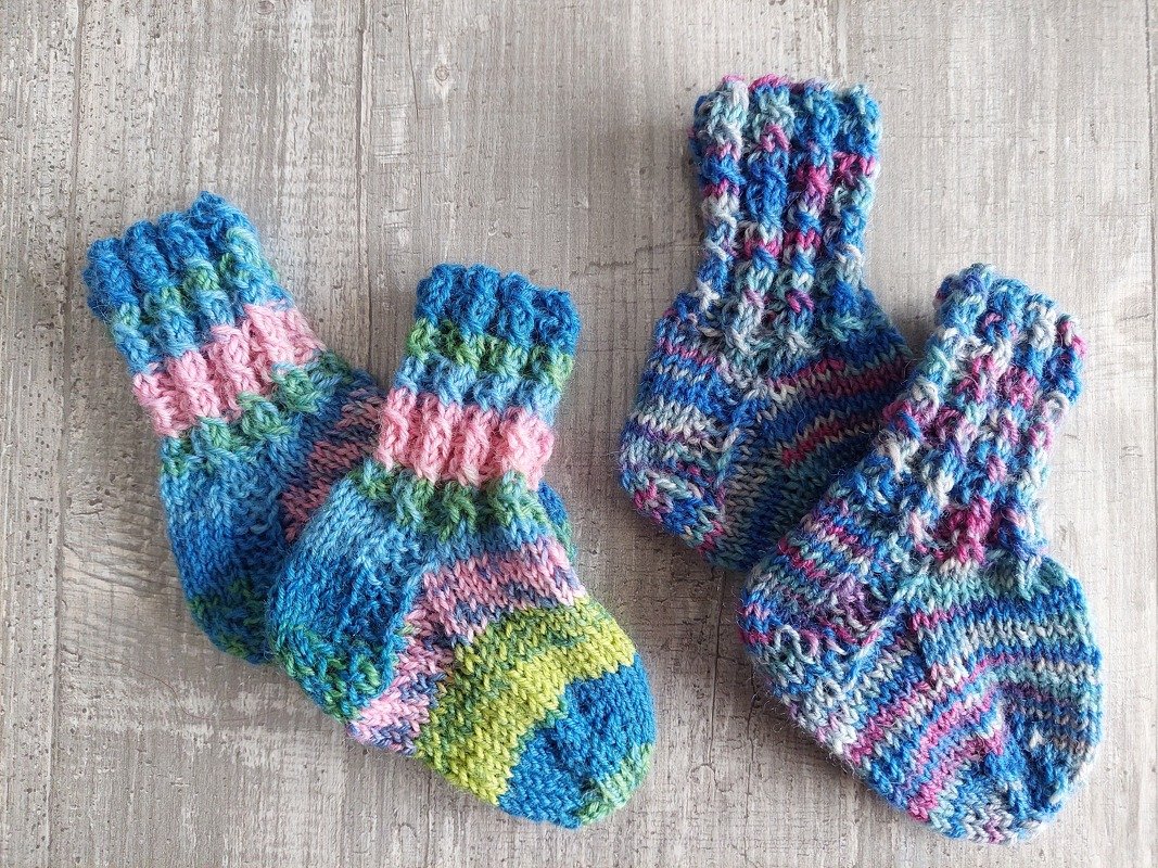 Zwei Paare bunter, handgestrickter Babysocken auf hellem Holzuntergrund, eines gestreift, das andere meliert.